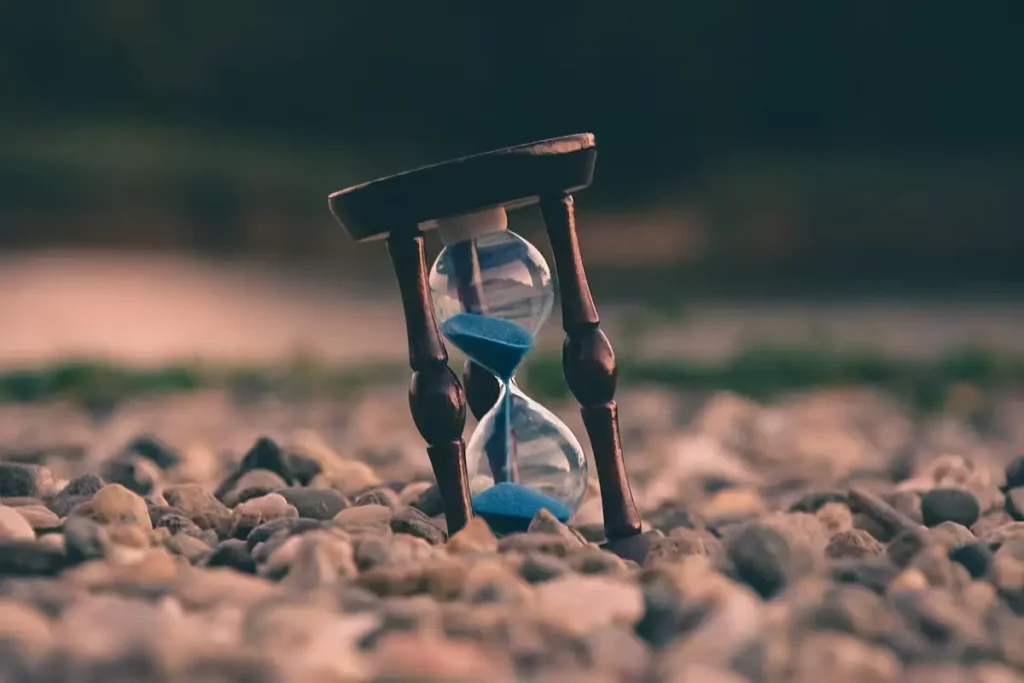 A small wooden hourglass with blue sand standing on a bed of pebbles